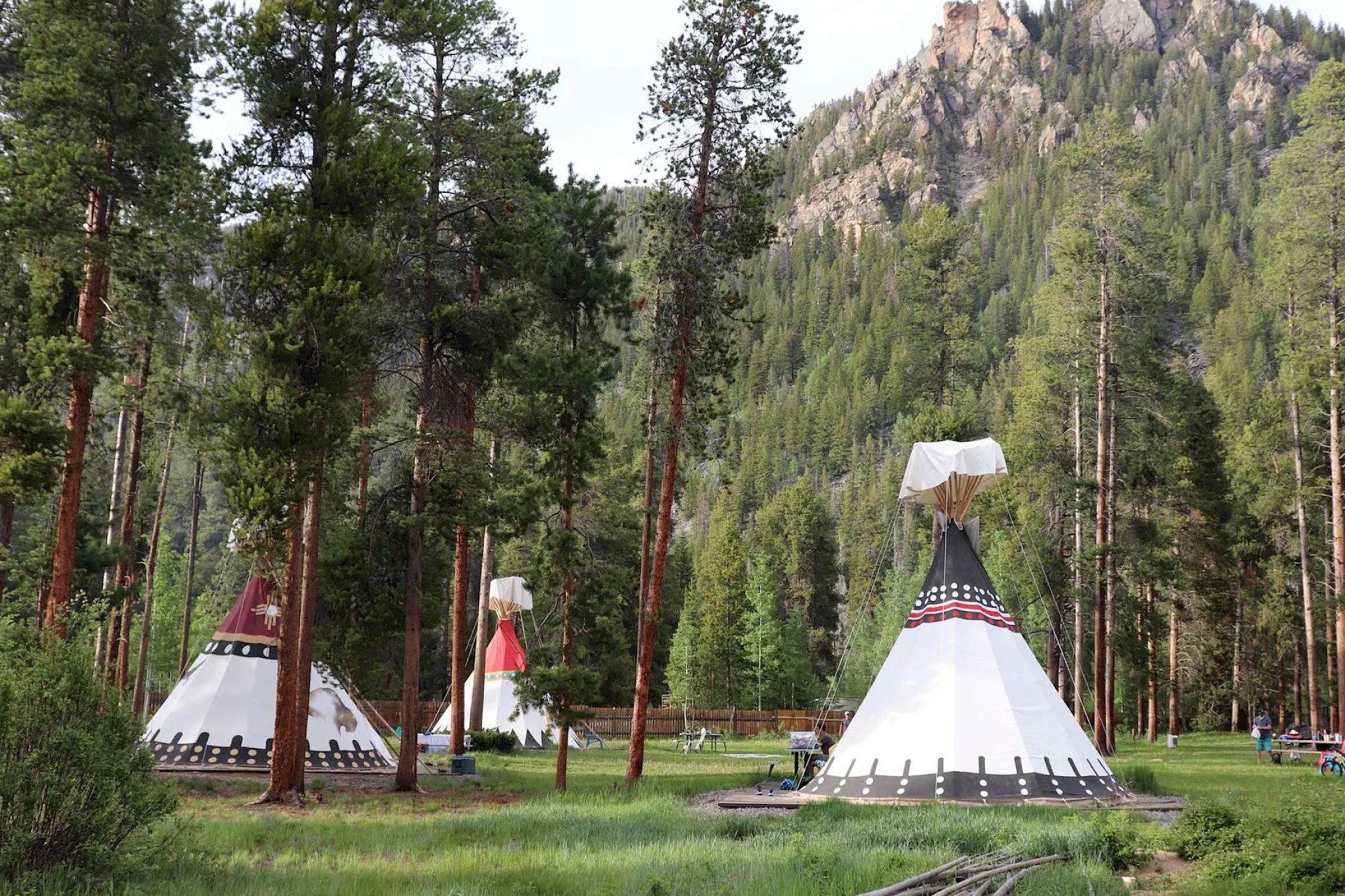 The image shows a scenic forested area with tipis set amid tall trees and mountains in the background.