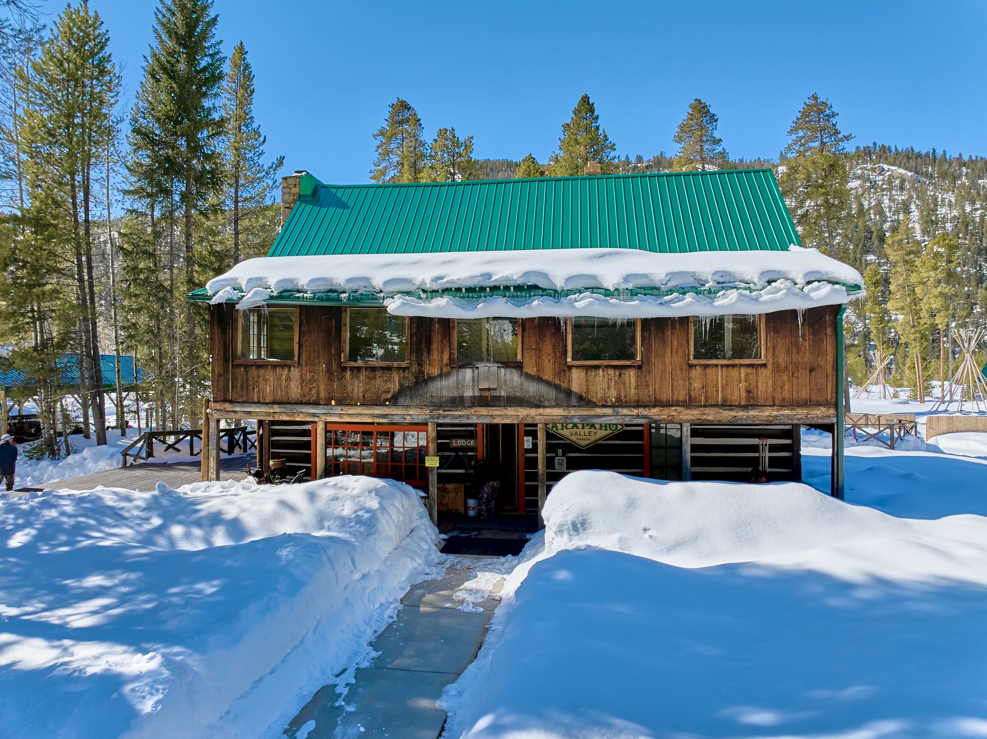 A wooden cabin with a green roof sits in a snowy forest, thick snow on the roof and ground, a clear blue sky above, wintery surroundings.
