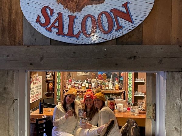 Three people in white outfits sit by the bar inside a rustic tavern under a &ldquo;Red Dog Saloon&rdquo; sign, sharing a casual moment.