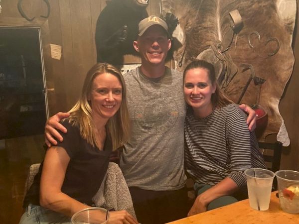 Three friends posing at a wooden bar, smiling with arms around each other in a cozy, rustic setting.