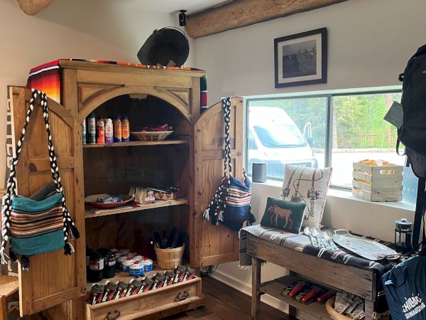 A rustic room with wooden beams, a tall armoire filled with clothes and items, a window bench, and a small desk cluttered with gear.