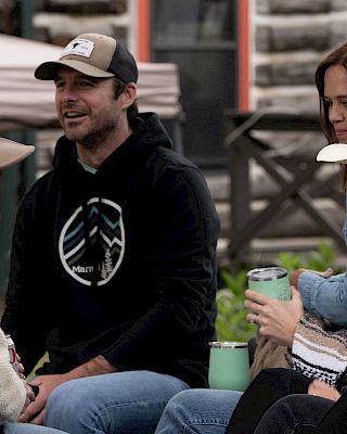 A group of four friends sit outdoors, chatting and smiling; one wears a wide-brim hat, another holds a drink, and a casual, relaxed vibe.