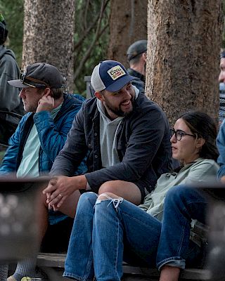 A group of friends sit near a campfire in a forest, chatting and relaxing by the flames as night begins.