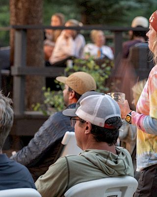 A group of casually dressed people sit outdoors on white chairs; a woman in a tie-dye shirt and knit hat stands nearby.