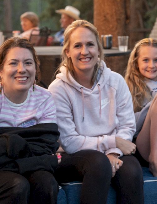 Three smiling women sit on a blue outdoor couch with drinks nearby, enjoying a sunset gathering.