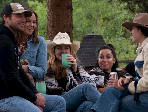 A group of friends sits outdoors around a campfire, smiling and chatting, some holding mugs; a casual hangout in a natural setting.