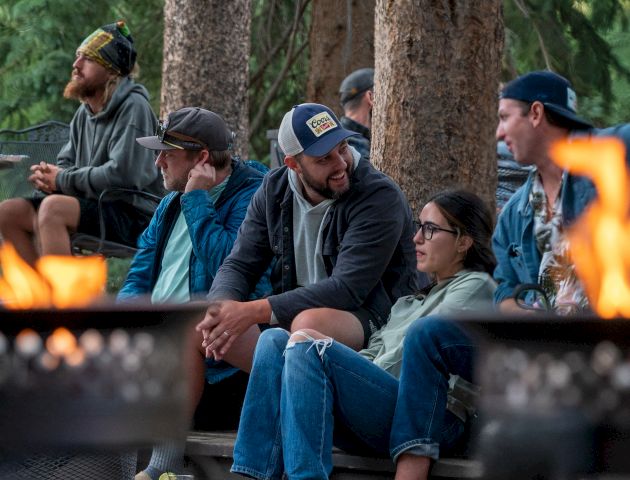 A group of friends sits by a campfire in a forest, chatting and listening, sunny outdoor gathering with flames in the foreground.