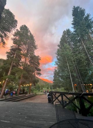 Sunset glow over a wooden boardwalk, tall pine trees frame a calm park path, people mingle near railings as orange-pink clouds drift overhead.